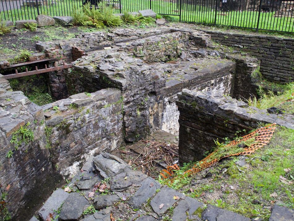 Brick walls defining pits in the ground, overgrown with grass.